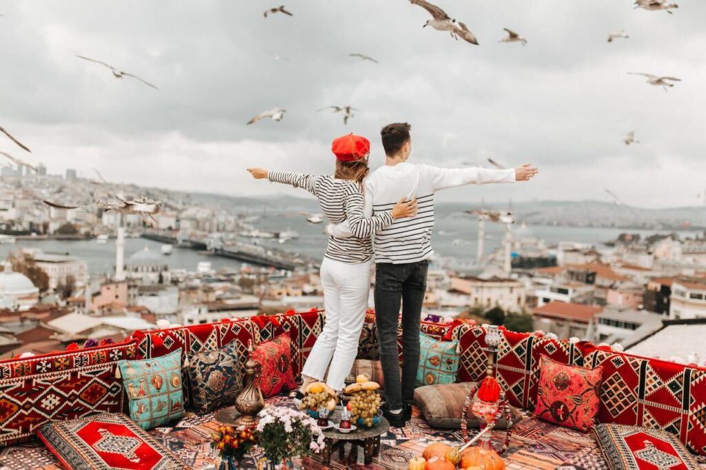 Couple standing on tippy toes arms raised in joy looking out over instanbul Turkey while surrounded by colorful fabrics and couches. Luxury Travel Destinations