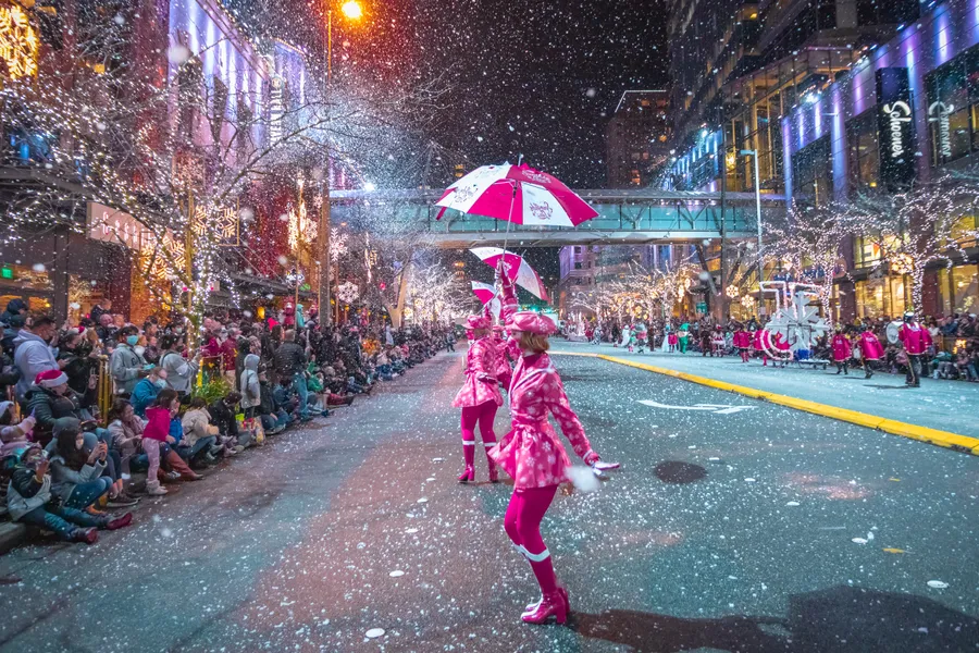 Seatle's Winterfest picture of a dancer dressed in winter dance attire holding pink umbrella with snow falling at night in a christmas light decorated street